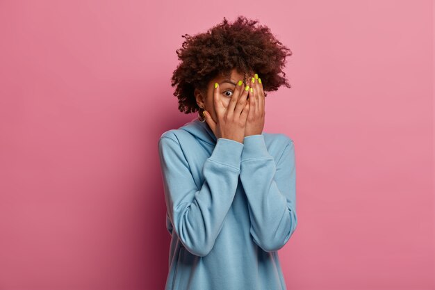 Die verängstigte Afroamerikanerin bedeckt das Gesicht mit Handflächen, starrt durch die Finger, hat Angst vor etwas, trägt ein blaues Sweatshirt, versteckt sich vor etwas Schrecklichem, das an einer rosa Wand isoliert ist