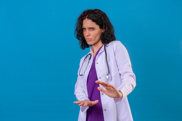 Die verängstigte junge Ärztin, die einen weißen Kittel mit einem Stethoskop trägt, hält ihre Hände hoch und sagt, dass sie auf isoliertem Blau nicht näher kommt