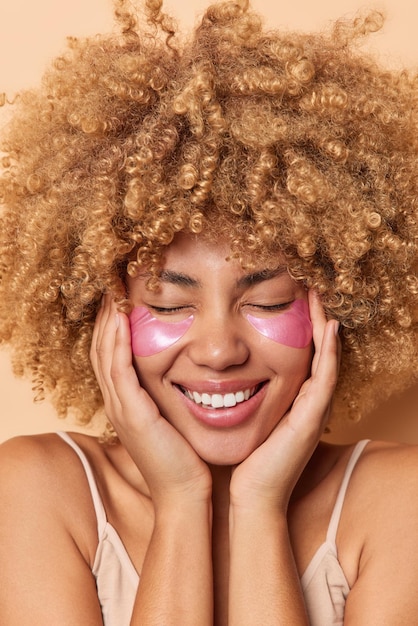 Die vertikale Aufnahme einer positiv gelockten Frau berührt das Gesicht, trägt sanft rosa Hydrogel-Patches unter den Augen auf, um Falten zu entfernen, lächelt fröhlich und hält die Augen geschlossen, steht nackten Schultern Beauty-Konzept