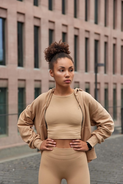 Die vertikale Aufnahme einer schlanken Frau mit lockigem Haar hält die Hände auf der Taille und trägt Sportkleidung, die sich auf Distanz konzentriert.