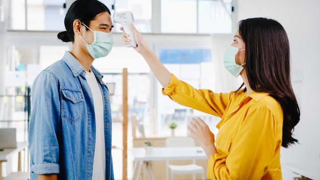 Die weibliche Empfangsdame in Asien, die eine tragende Gesichtsmaske trägt, verwendet vor dem Betreten des Büros einen Infrarot-Thermometerprüfer oder eine Temperaturpistole auf der Stirn des Kunden. Lebensstil neu normal nach Koronavirus.