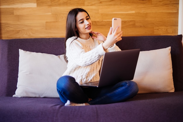 Die zarte Dame arbeitet an einem Laptop und telefoniert auf dem Bett im Haus