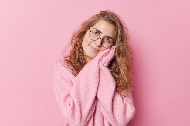 Die zarte, sanfte junge Europäerin hält die Hände in der Nähe des Gesichts, lächelt glücklich und trägt eine runde Brille und einen Kaschmirpullover isoliert auf rosa Hintergrund. Lächelndes langhaariges weibliches Modell starrt Kamera an