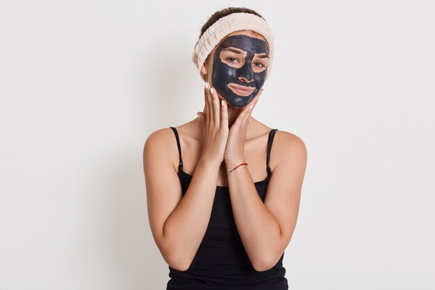 Die zufriedene europäische Frau trägt eine pflegende Tonmaske auf das Gesicht auf, hat einen fröhlichen Ausdruck, berührt die Wangen und hat Probleme mit trockener Haut.