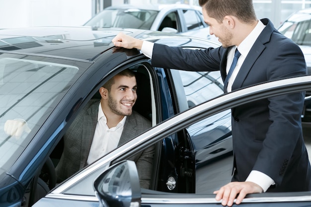 Dies ist das eine horizontale Porträt eines gutaussehenden jungen Geschäftsmannes, der im Auto sitzt und mit einem Autohändler spricht, der in der Nähe steht