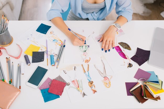 Dieses Bild beschreibt die Prozesse beim Entwerfen von Kleidung. Es gibt Hände eines Mädchens, die Skizzen auf den Tisch zeichnen. Es gibt kreatives Durcheinander mit verschiedenen Dingen auf dem Tisch.
