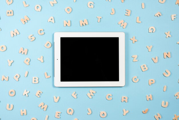 Digital-Tablette mit der schwarzen Bildschirmanzeige umgeben mit hölzernen Buchstaben auf blauem Hintergrund