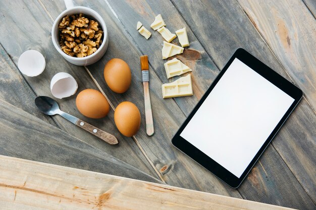 Digitale Tablette in der Nähe von Schokoladenstücken; Eier und Walnüsse auf hölzernen Hintergrund