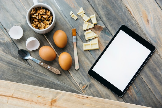 Digitale Tablette in der Nähe von Schokoladenstücken; Eier und Walnüsse auf hölzernen Hintergrund