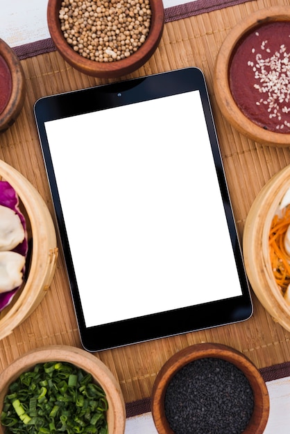 Digitales Tablett mit weißem leerem Bildschirm umgeben mit Dampfern; Koriandersamen; Sesam und Frühlingszwiebeln auf Tischset