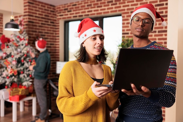 Diverse Kollegen, die im festlichen Büro auf den Computer schauen und während der Winterferienzeit in einem mit Weihnachtsschmuck und Lichtern geschmückten Raum arbeiten. Menschen mit Weihnachtsmütze bei der Arbeit am Heiligabend.