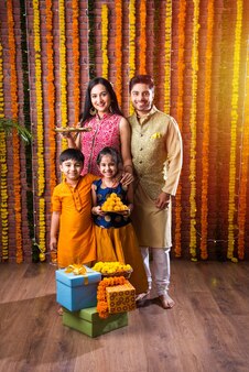 Diwali- oder rakshabandhan-feier - indische junge vierköpfige familie, die das deepavali- oder bhai-dooj-festival mit süßem laddoo, öllampe oder diya und geschenkboxen feiert, essen isst oder selfies macht