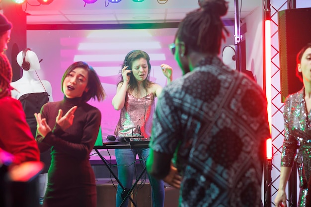 DJ-Frau mit Kopfhörern tritt mit einer Mischstation für elektronische Musik auf der Bühne auf, während sie im Nachtclub tanzt. Menschen feiern auf der Tanzfläche und gehen in die Diskothek