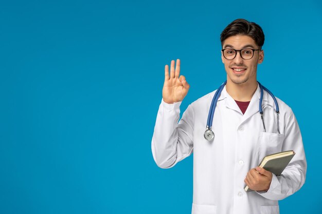Doctors Day süßer junger brünetter Typ im Laborkittel mit Brille, der ein Ok-Zeichen zeigt und ein Buch hält