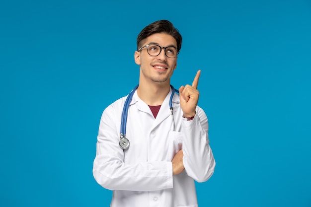 Doctors Day süßer junger brünetter Typ im Laborkittel mit Brille, der nachdenkt und nach oben schaut