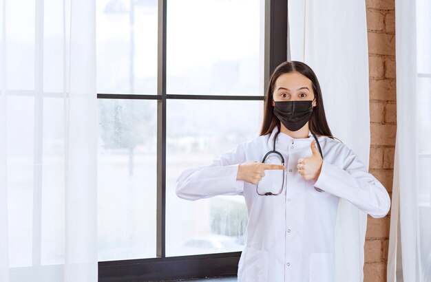 Doktor in der schwarzen Maske mit einem Stethoskop, das am Fenster steht und Zufriedenheitszeichen zeigt.