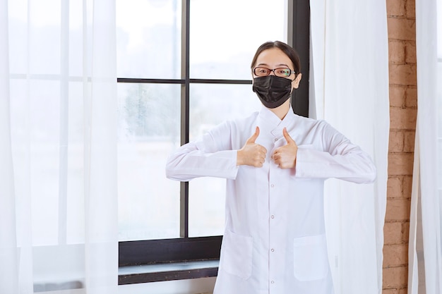 Doktor in der schwarzen Maske und in den Brillen, die am Fenster stehen und Genusszeichen zeigen.