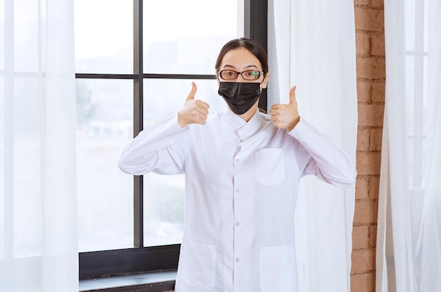 Doktor in der schwarzen Maske und in den Brillen, die am Fenster stehen und Genusszeichen zeigen.