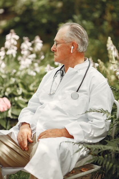Doktor in weißer Uniform. Alter Mann, der in einem Sommerpark sitzt. Senior mit Stethoskop.
