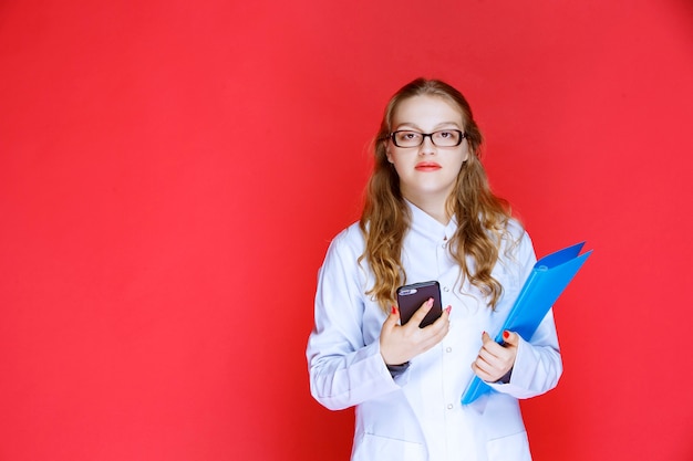 Doktor mit einem blauen Ordner und einer Brille, die ihr Telefon überprüft.