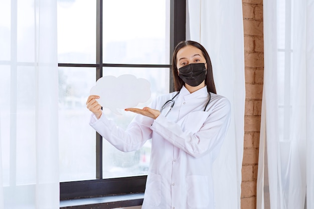 Doktor mit Stethoskop und schwarzer Maske, die ein Denkbrett der weißen Wolkenform halten.