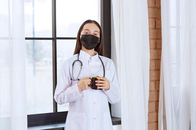 Doktor mit Stethoskop und schwarzer Maske hält eine schwarze Tasse Getränk in der Pause und wärmt ihre Hände.