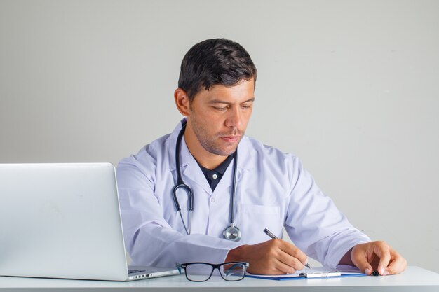 Doktor sitzt und macht sich Notizen im weißen Kittel und Stethoskop