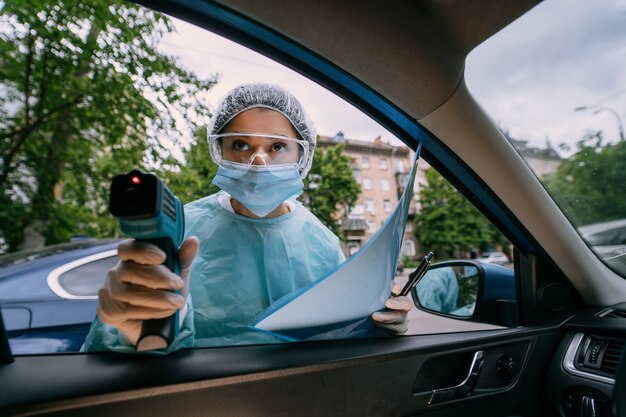 Doktorfrau verwenden Infrarot-Stirn-Thermometer-Pistole, um die Körpertemperatur zu überprüfen. Für Virus-Covid-19-Symptome. Frau mit dem Isolationskleid oder den Schutzanzügen und den chirurgischen Gesichtsmasken im Freien.