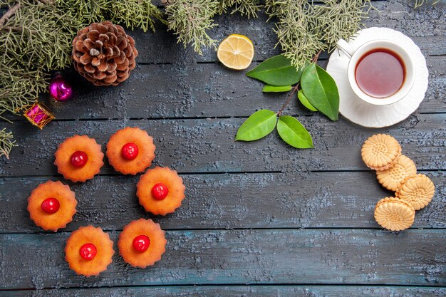 Draufsicht abgerundete Kirschcupcakes Kegeltannenbaum verlässt Weihnachtsspielzeugscheibe Zitronenscheibe eine Tasse Tee und Kekse auf dunklem Holztisch mit Kopierraum