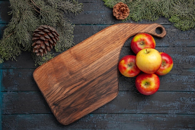 Draufsicht Apfel und Brett fünf gelb-rötliche Äpfel neben dem Holzschneidebrett auf grauem Tisch zwischen Ästen mit Zapfen