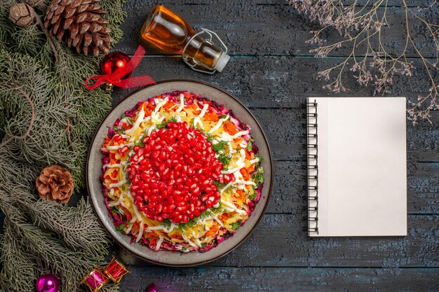 Draufsicht appetitliches Gericht Weihnachtsgericht mit Granatapfelkernen neben den weißen Notebook-Baumzweigen und Öl in der Flasche