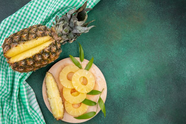 Draufsicht auf Ananas mit einem Stück aus ganzen Früchten und Ananasscheiben auf Schneidebrett auf kariertem Stoff und grüner Oberfläche ausgeschnitten