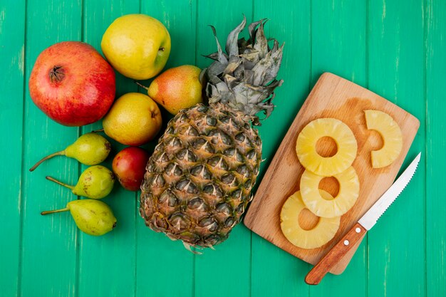 Draufsicht auf Ananasscheiben mit Messer auf Schneidebrett und Ananas-Granatapfel-Pfirsichpflaume auf grüner Oberfläche