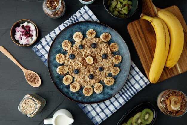 Draufsicht auf Banane Blackthorn Flachs Haferflocken in Teller auf kariertem Tuch mit Bananen-Walnuss-Smoothie im Glas und Banane Kiwi Hüttenkäse Milch Hafer und Leinsamen Walnuss auf schwarzem Hintergrund