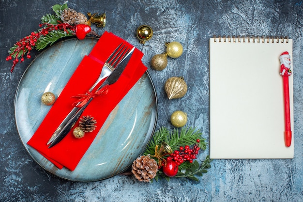 Draufsicht auf Besteck mit rotem Band auf einer dekorativen Serviette auf blauem Teller und Weihnachtszubehör neben Notizbuch mit Stift auf dunklem Hintergrund