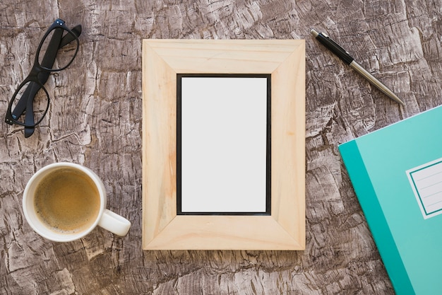 Draufsicht auf Bilderrahmen; Kaffeetasse; Brille; Stift und Notizbuch auf strukturiertem Hintergrund