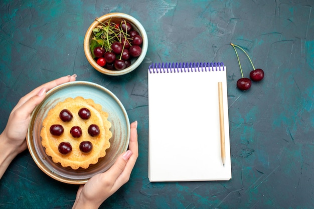 Draufsicht auf Cupcake mit Kirschen, die jemand neben Notizbuch hält