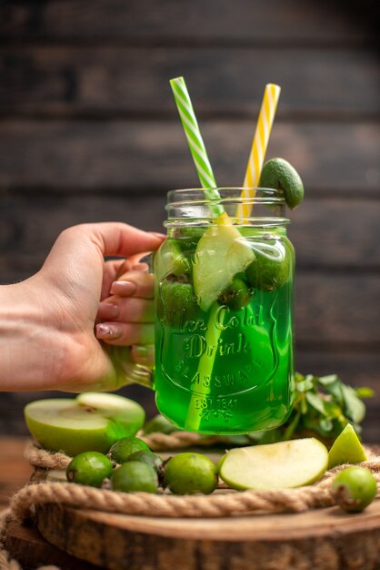 Draufsicht auf die Hand, die ein Glas mit frischem, köstlichem Fruchtsaft hält, serviert mit Apfel und Feijoas auf einem Holzbrett auf einem braunen Tisch