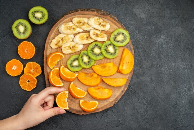 Draufsicht auf die Hand, die eine Orangenscheibe aus einem natürlichen organischen frischen Obst auf einem Schneidebrett auf dunklem Hintergrund nimmt