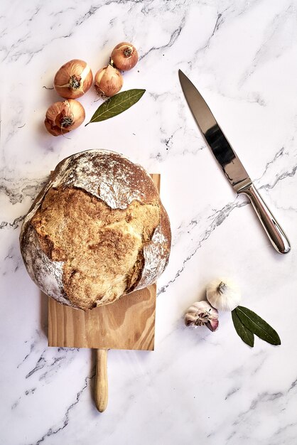 Draufsicht auf ein frisch gebackenes traditionelles Brot auf einem Holzbrett mit Zwiebeln, Knoblauch und einem Messer