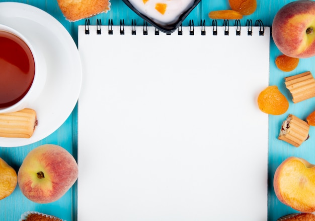 Draufsicht auf ein Skizzenbuch und frische Pfirsiche mit Muffins getrockneten Aprikosenkeksen und einer Tasse Tee, die auf Blau angeordnet sind