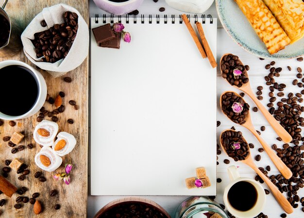 Draufsicht auf ein Skizzenbuch und Kaffeebohnen in der Holzschale und in den Löffeln und mit lokum Schokoladenstücken und Kaffeebohnen, die auf rustikalem Hintergrund verstreut sind