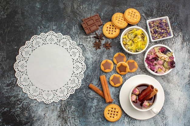 Draufsicht auf ein Stück weiße Spitze mit Kräutertee und Keks und trockenen Blumen auf grauem Grund