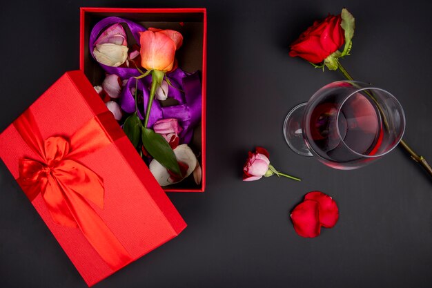 Draufsicht auf eine offene rote Geschenkbox mit Rosenblume und einem Glas Rotwein auf schwarzem Tisch