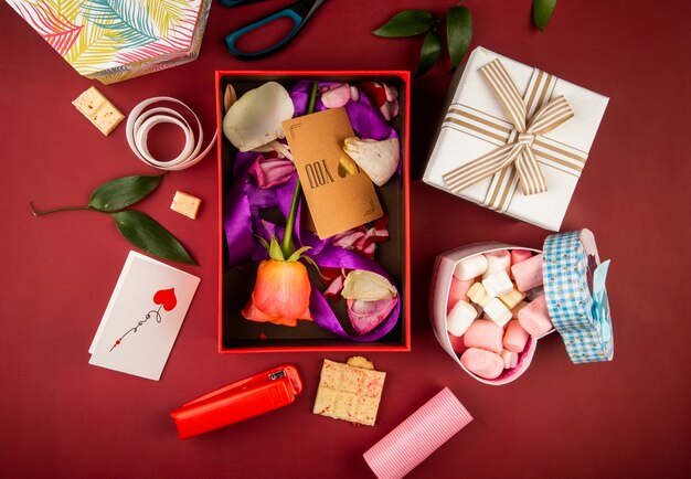 Draufsicht auf eine rote Geschenkbox mit brauner Papierkarte und korallenfarbener Rosenblume und Blütenblättern mit lila Band und herzförmiger Box, gefüllt mit Marshmallow auf dunkelrotem Tisch