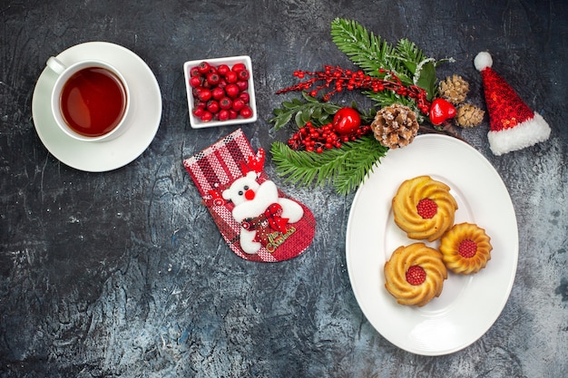 Kostenloses Foto draufsicht auf eine tasse tee köstliche kekse auf einem weißen teller weihnachtsmann-hut und schokolade in einer schüssel auf dunkler oberfläche