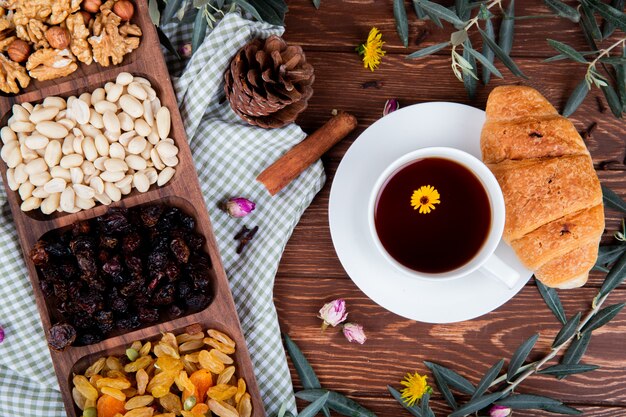 Draufsicht auf eine Tasse Tee mit Croissant, gemischten Nüssen mit getrockneten Früchten und verstreutem Löwenzahn auf Holz