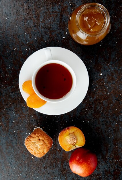 Draufsicht auf eine Tasse Tee mit frischem süßem Nektarinenmuffin und einem Glas mit Pfirsichmarmelade auf Schwarz