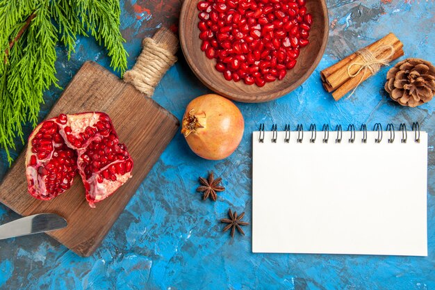 Draufsicht auf einen geschnittenen Granatapfel und ein Tafelmesser auf Schneidebrett Granatapfelkerne in Schüssel und Granatäpfel Zimt-Anis-Samen ein Notizbuch auf blauem Hintergrund