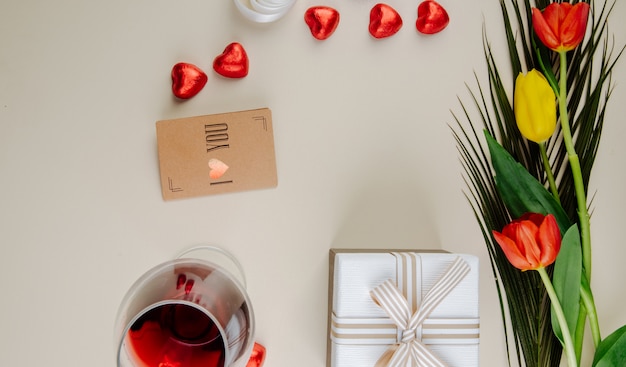 Draufsicht auf einen Strauß Tulpen mit herzförmigen Pralinen, eingewickelt in rote Folie, Glas Wein, kleine braune Papiergrußkarte und eine Geschenkbox auf weißem Tisch